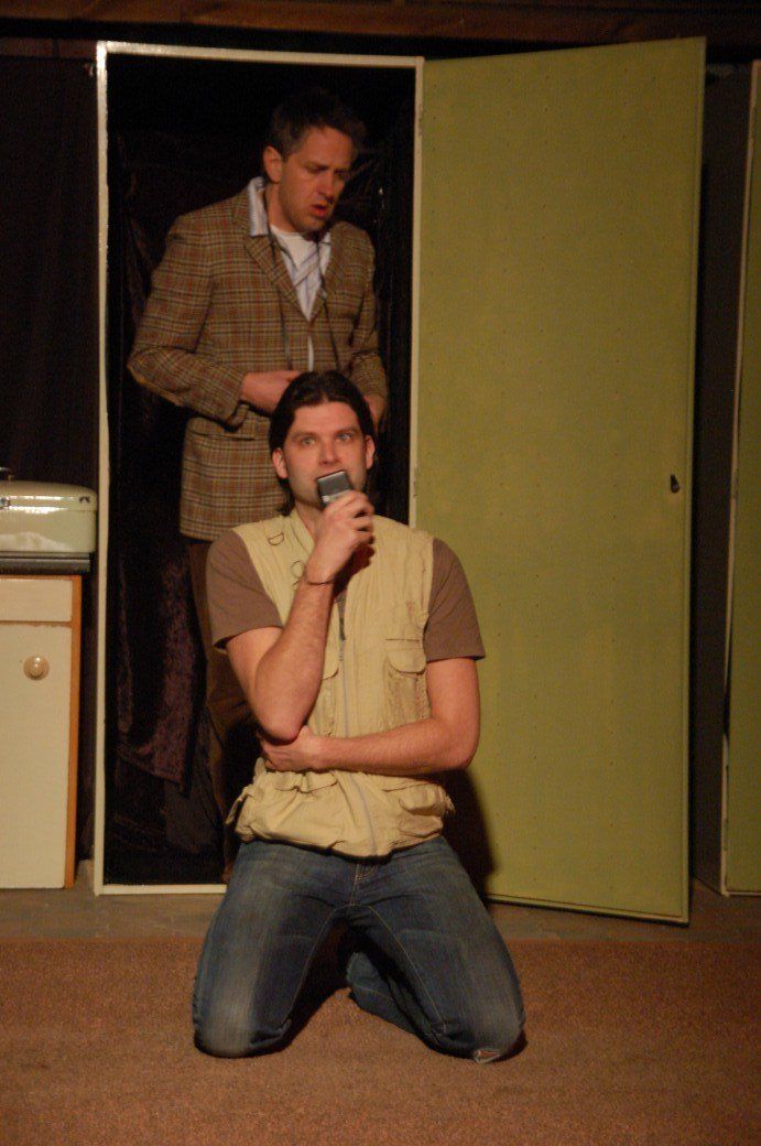Man kneeling and speaking into a microphone on a stage, with another man standing in a doorway behind him.