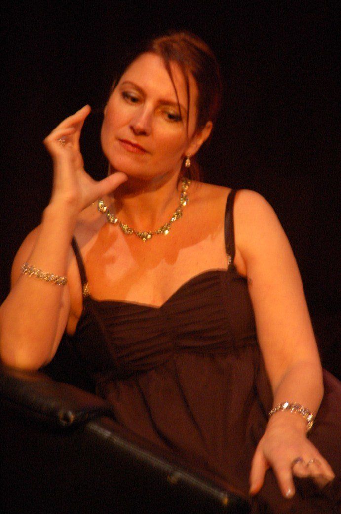 Woman in a black dress posing against a dark background, hand raised near her face.
