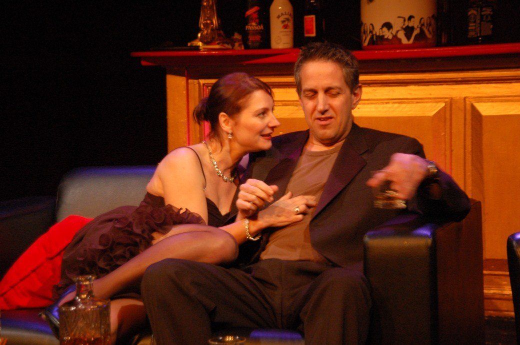 Two people sitting on a couch in a warmly lit room, talking closely while holding drinks
