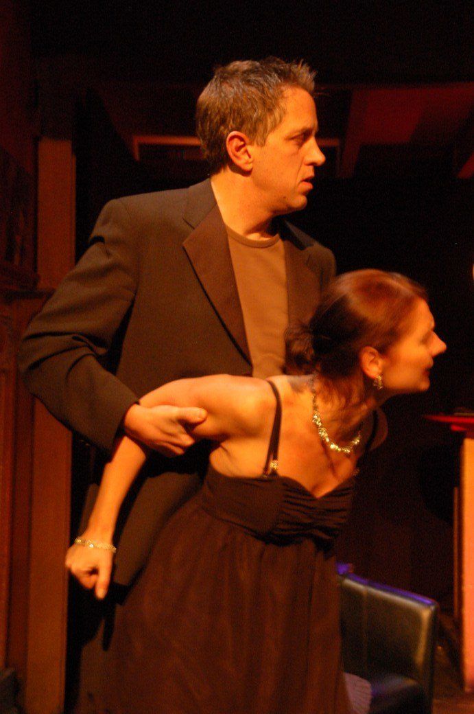 Two performers on a dimly lit stage, one in a dark dress leaning forward while another stands behind her.