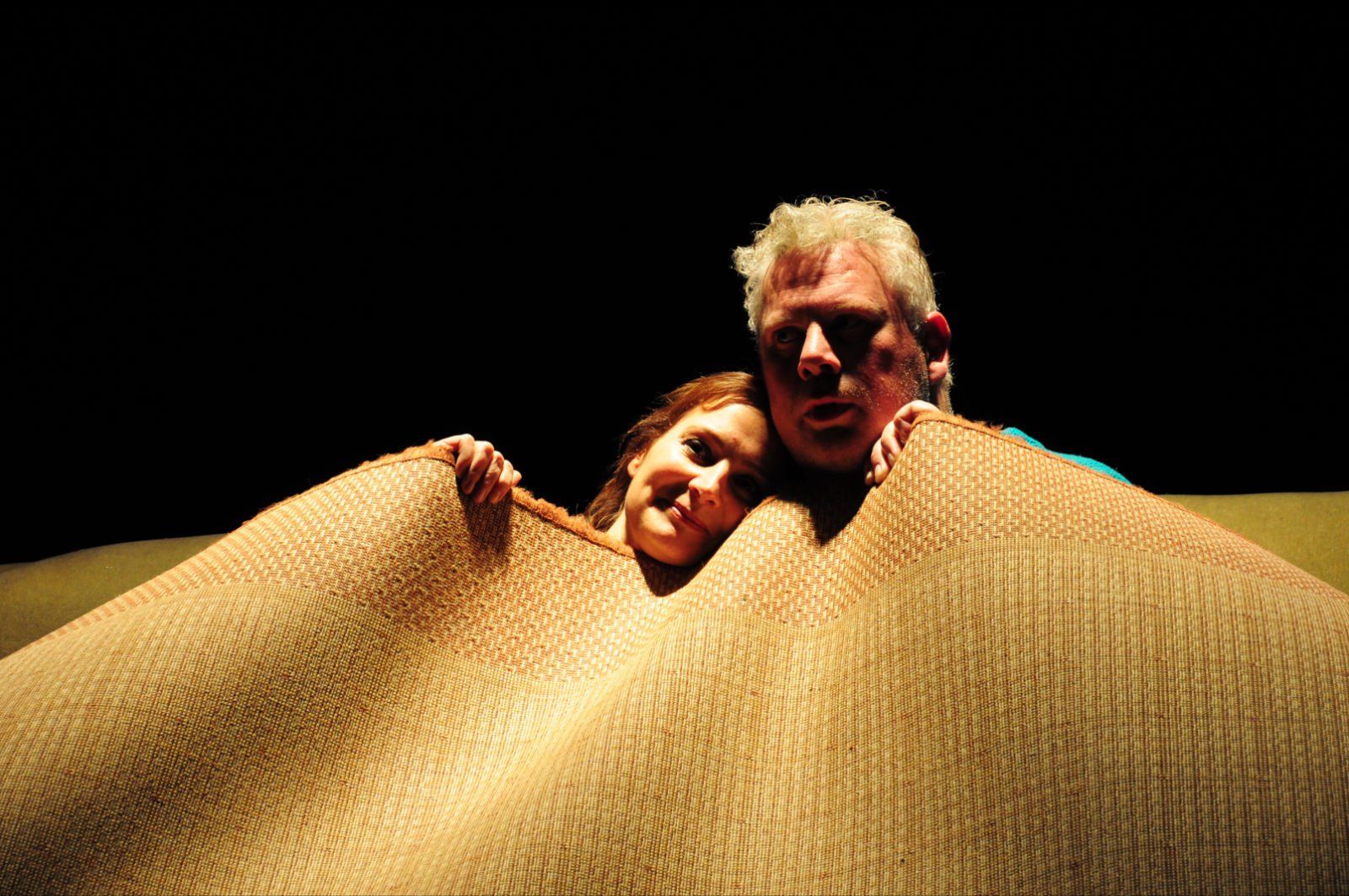 Two people peeking over a large tan woven blanket against a black background