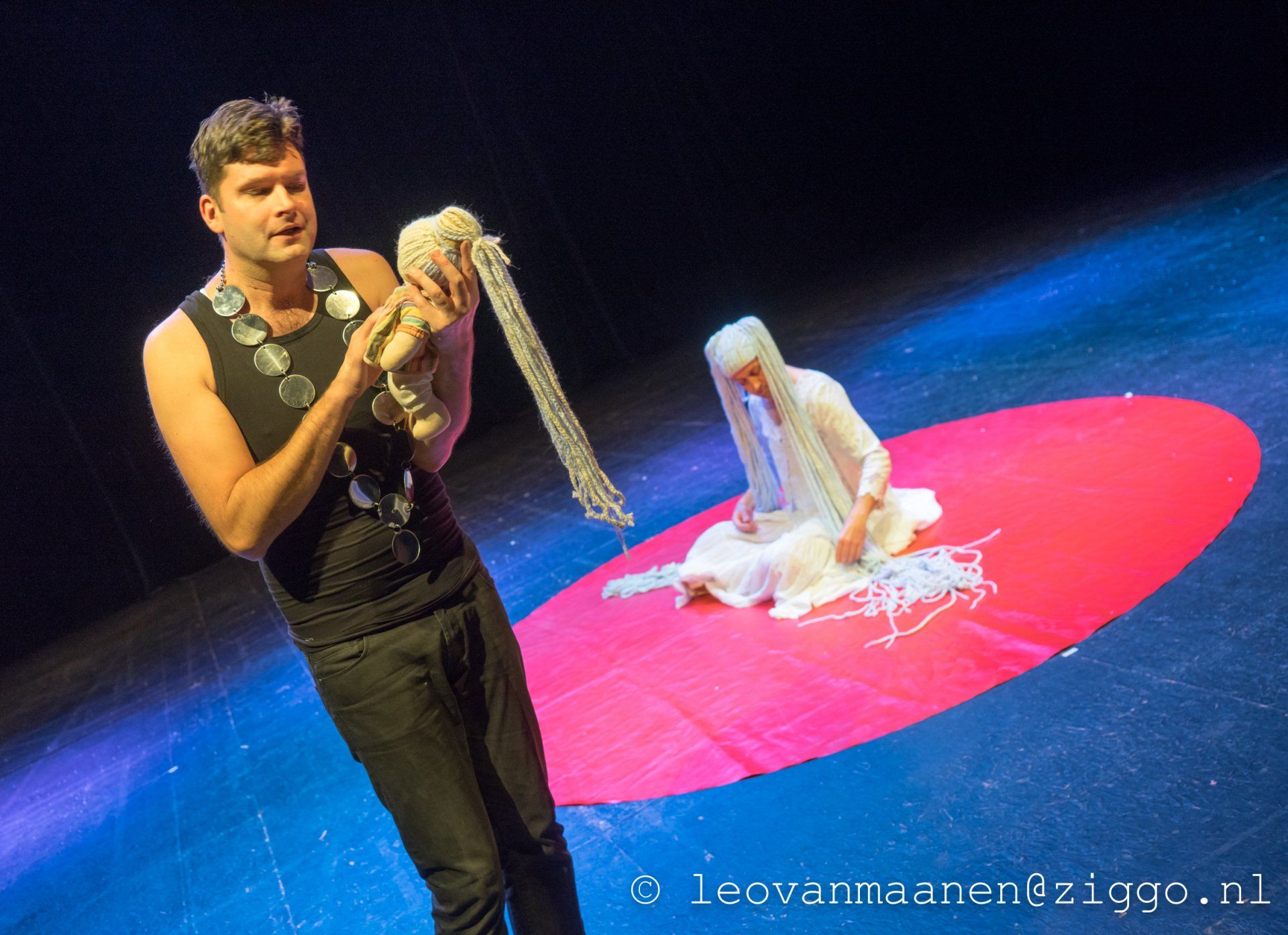 Performers on a stage with a bright red circle and blue-lit floor, one holding a long white prop.