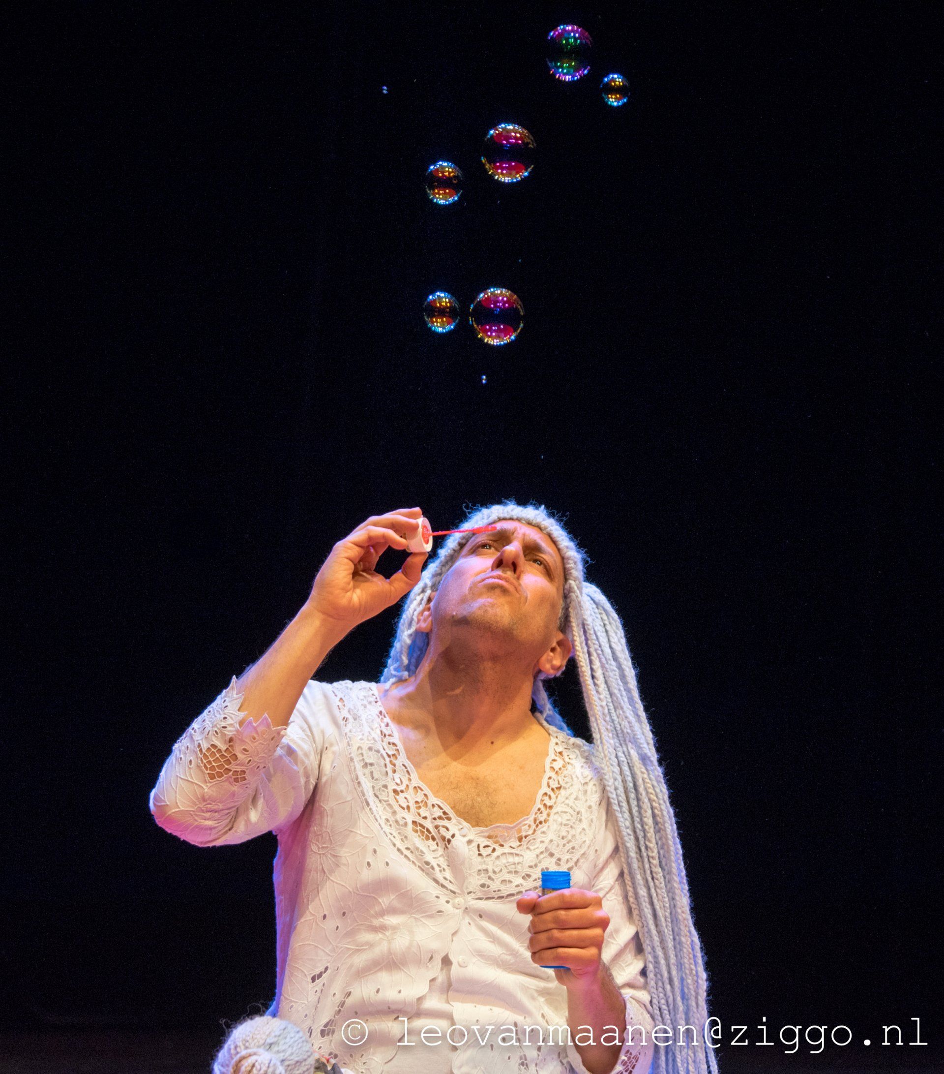 Woman in white blowing soap bubbles on a dark stage, holding a small bottle.