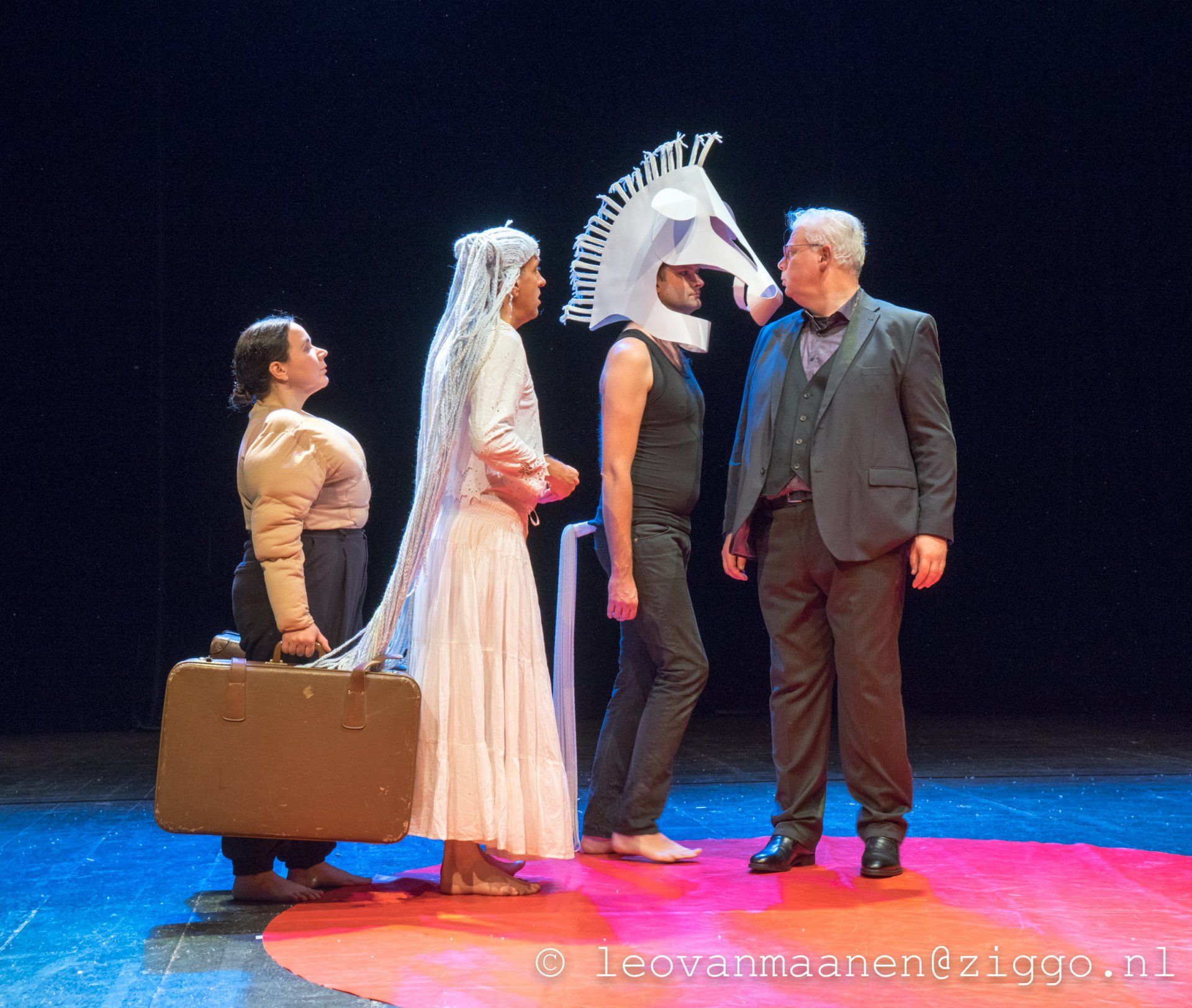 Four people on a stage, one carrying a suitcase, another wearing a white dress and feathered headpiece.