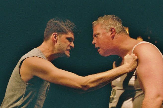 Two men in sleeveless shirts face off intensely against a dark background, one gripping the other’s shoulder.