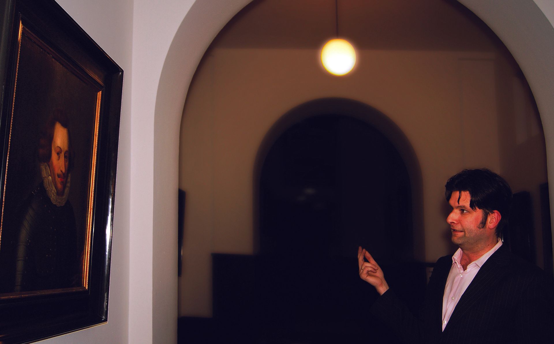 Man in a dark suit gestures beside a framed portrait in a dim arched gallery hallway