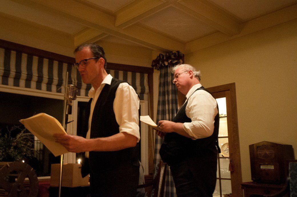 Two men in vests reading from papers at microphones in a warmly lit room.