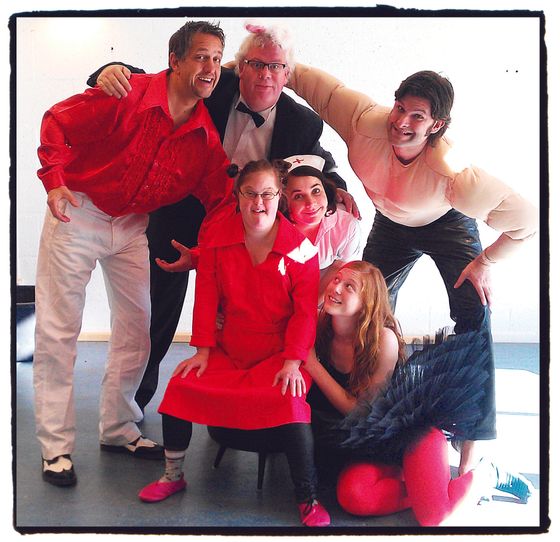 Six people pose together indoors, smiling and leaning into a group huddle.