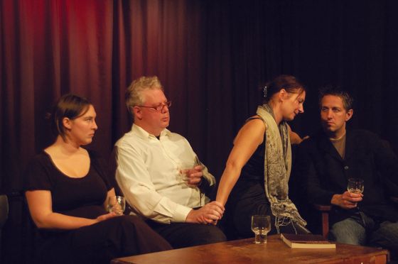 Four people seated on a stage, conversing under red curtains beside a small table with glasses.