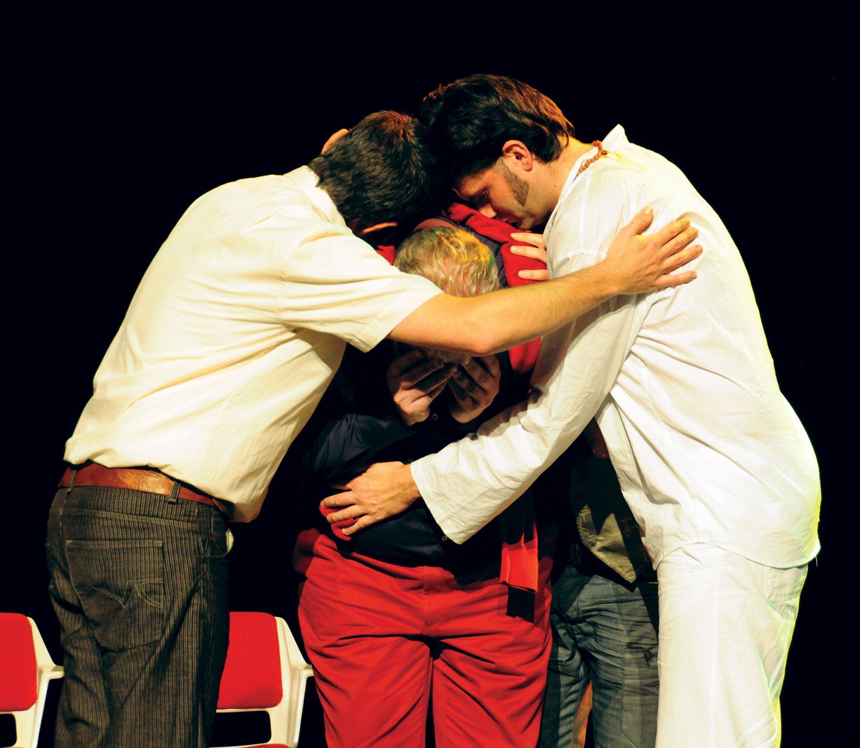 Three people embracing in a group hug on stage, wearing white and red clothing