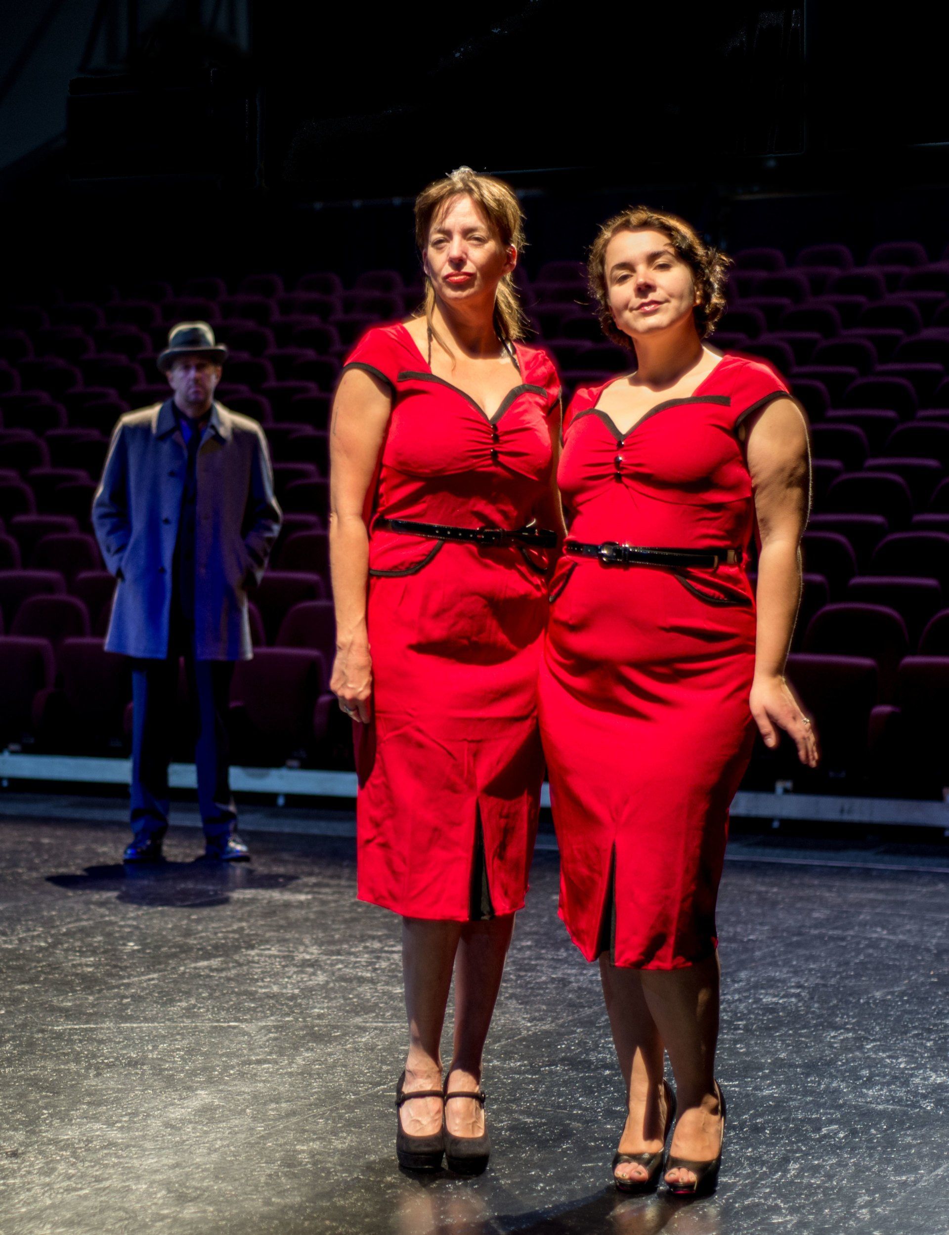 Two women in matching red dresses pose on a theater stage, with a man in a blue coat behind them.