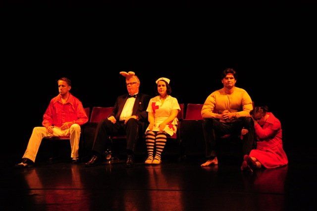 Five performers seated on a dark stage under warm red-orange lighting