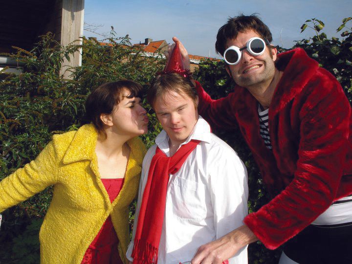 Three people posing outdoors in colorful costumes, including a white coat, red scarf, and red jacket with goggles
