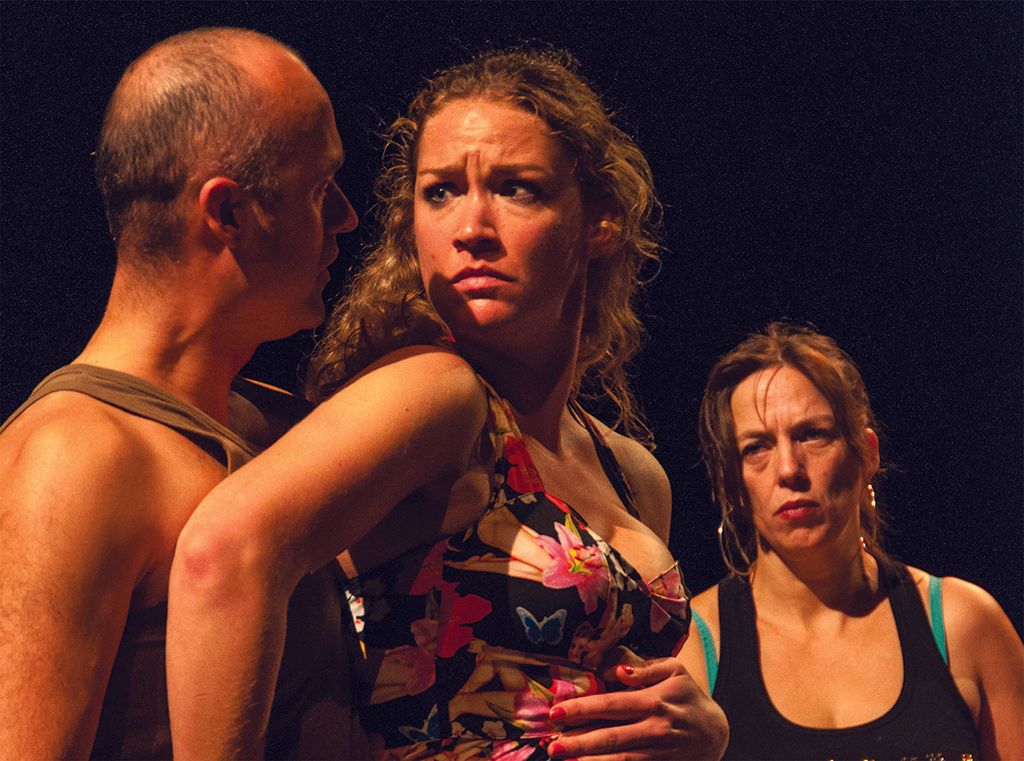 Three dancers in dramatic stage lighting, with a tense woman in a floral top at center.