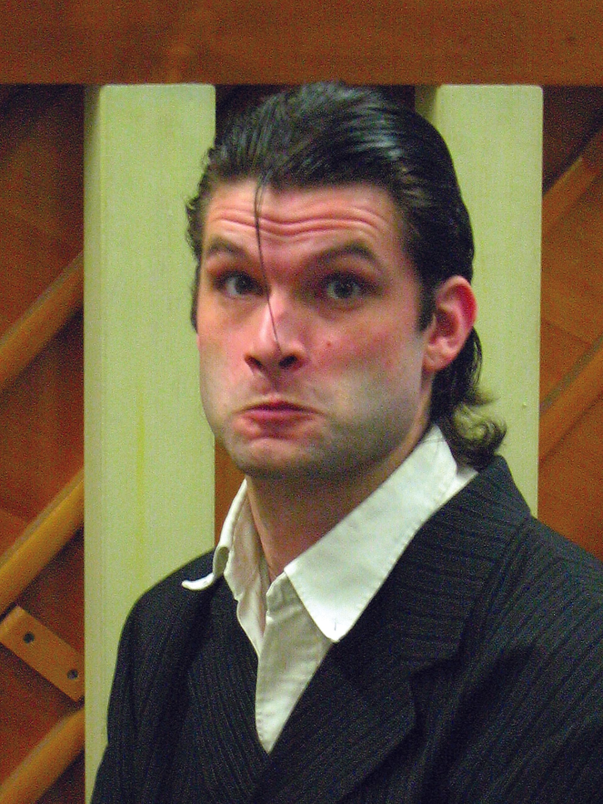 Man in a dark suit and white shirt making a puckered expression indoors against a yellow background