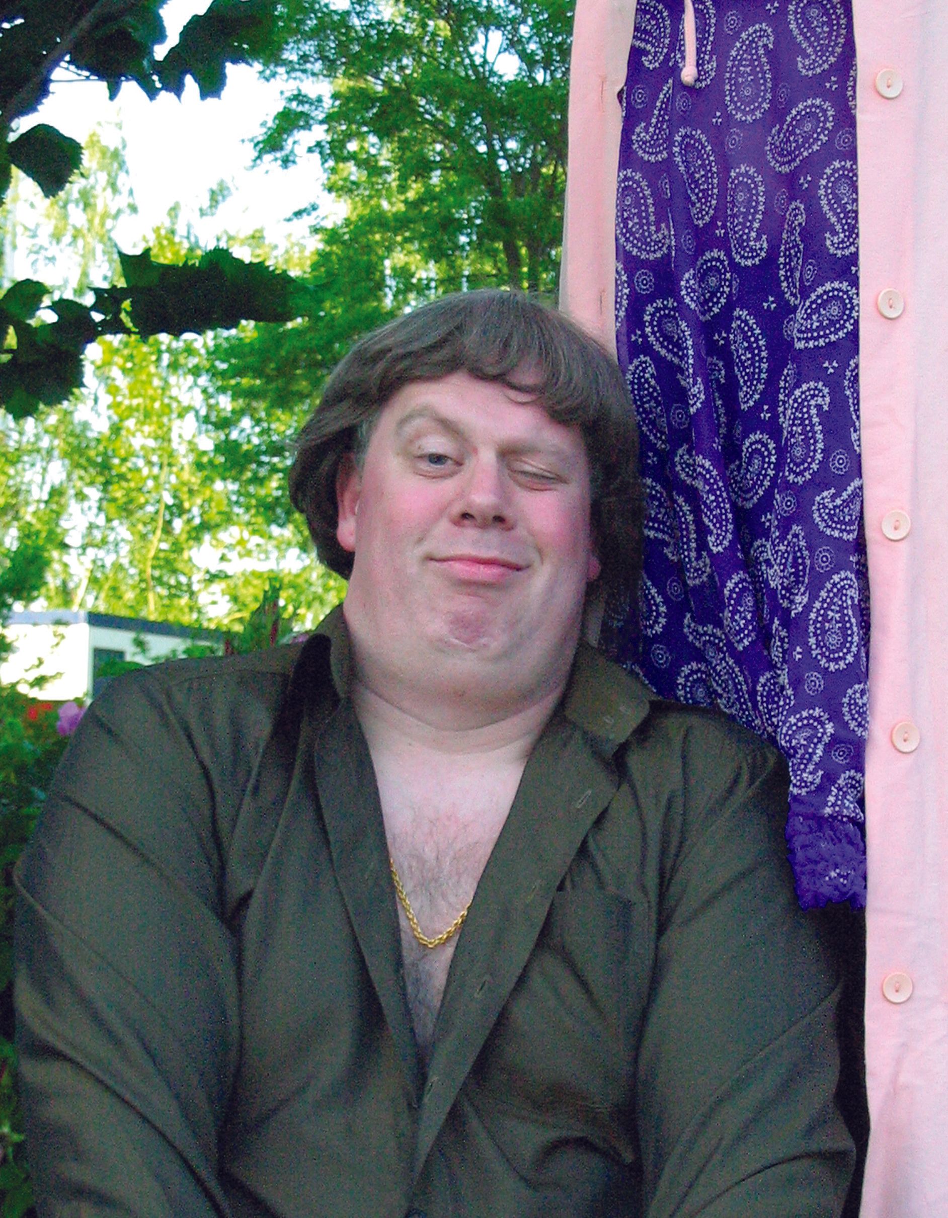 Person in a dark jacket seated outdoors beside a blue patterned curtain and pink post, with a slight smile.