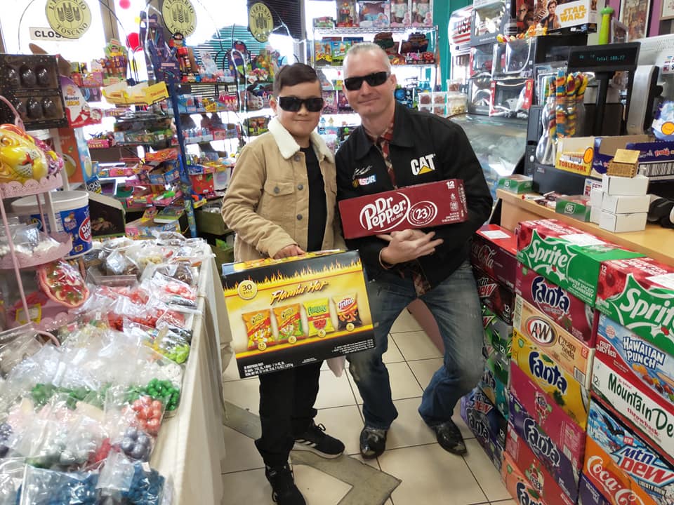 A person in a tan jacket and an adult in a black CAT jacket hold snacks and soda inside a shop with shelves of candy.