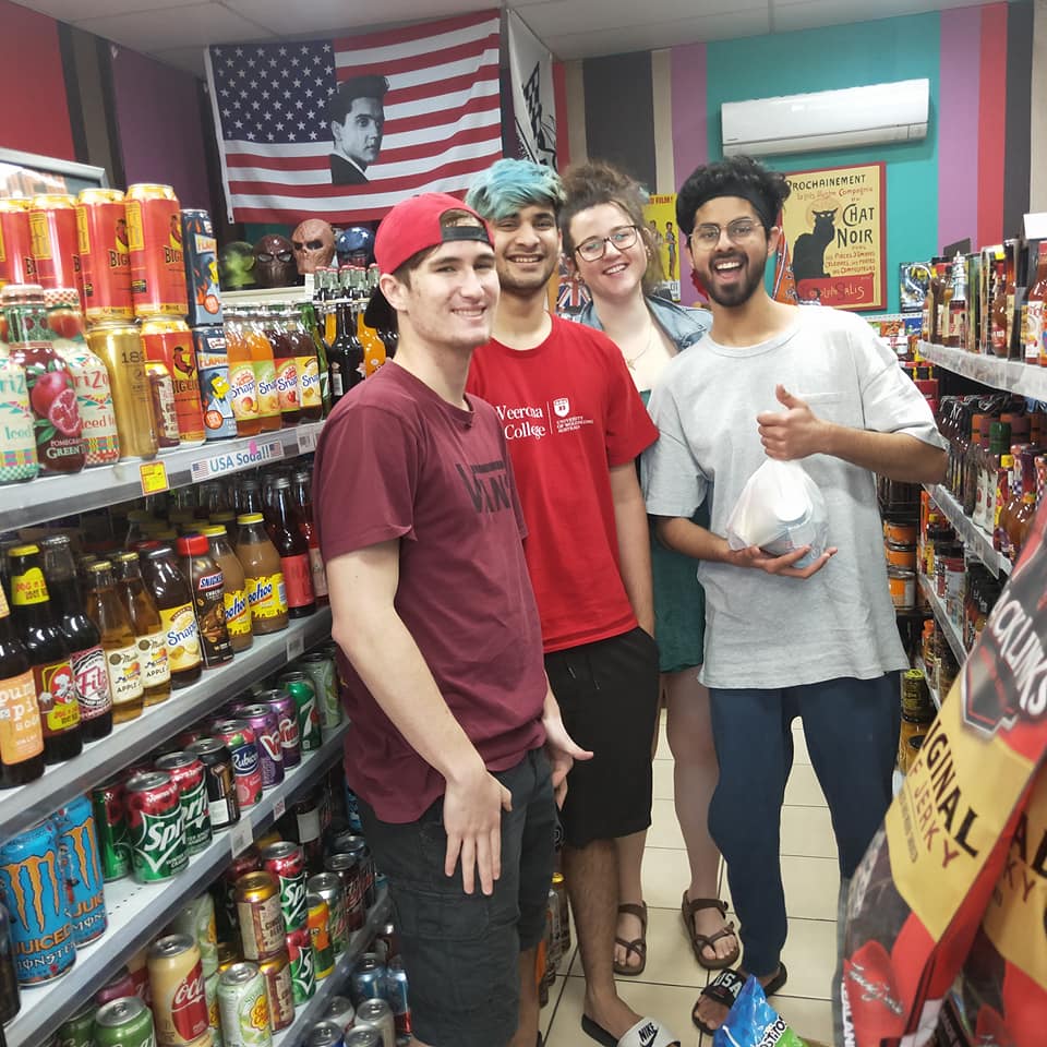 Four smiling people stand in a convenience store aisle filled with shelves of beverages, posing for a group photo.