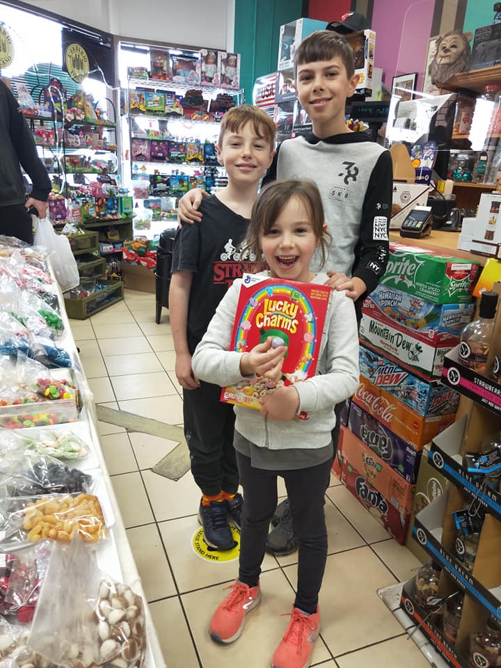 Three smiling children stand in a shop, with the girl in front holding a box of Lucky Charms cereal.