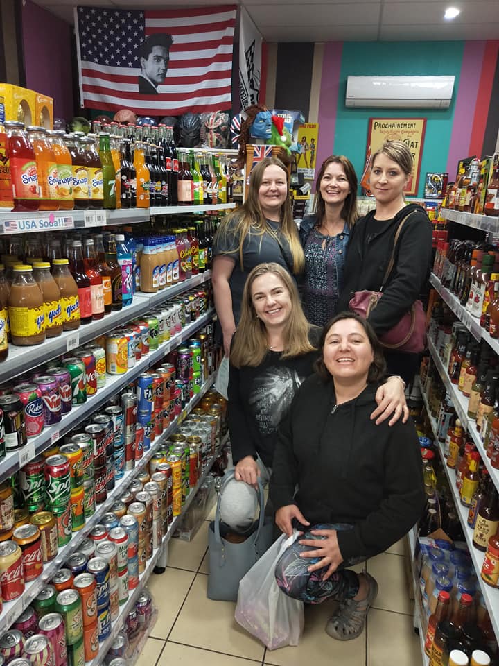 Five smiling people pose in the aisle of a store filled with shelves of beverages, with a US flag hanging in the background.