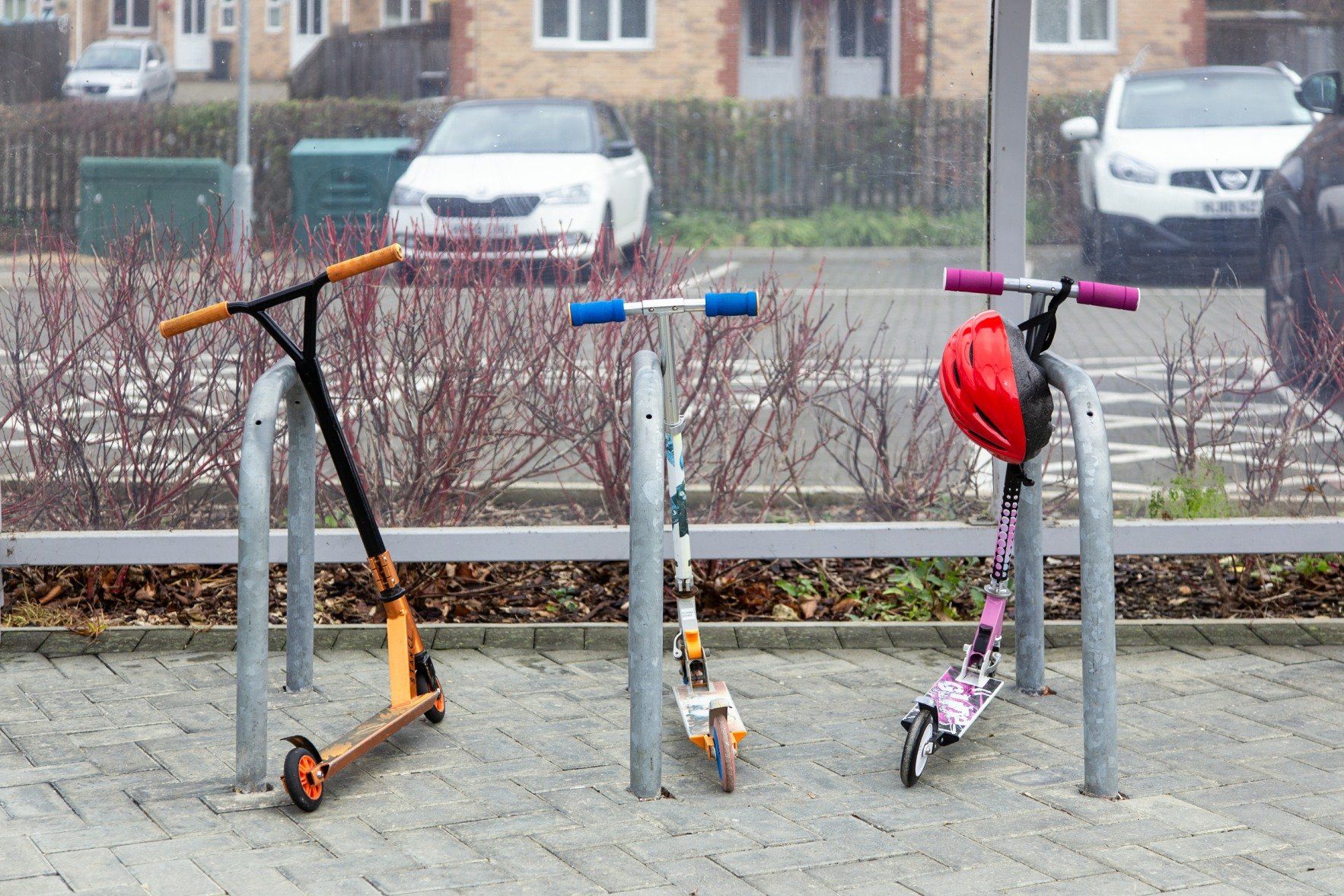 Three scooters parked in a metal rack; orange, white/yellow, and pink/black, with a red helmet on the last one.