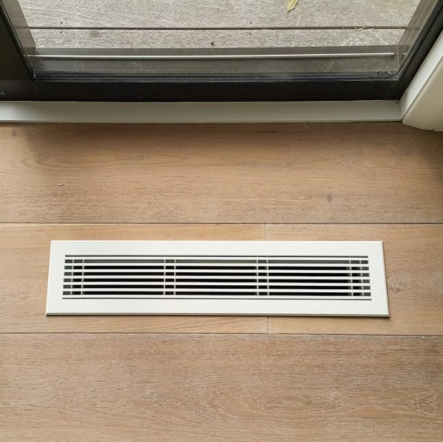 A white air vent is sitting on a wooden floor next to a window.