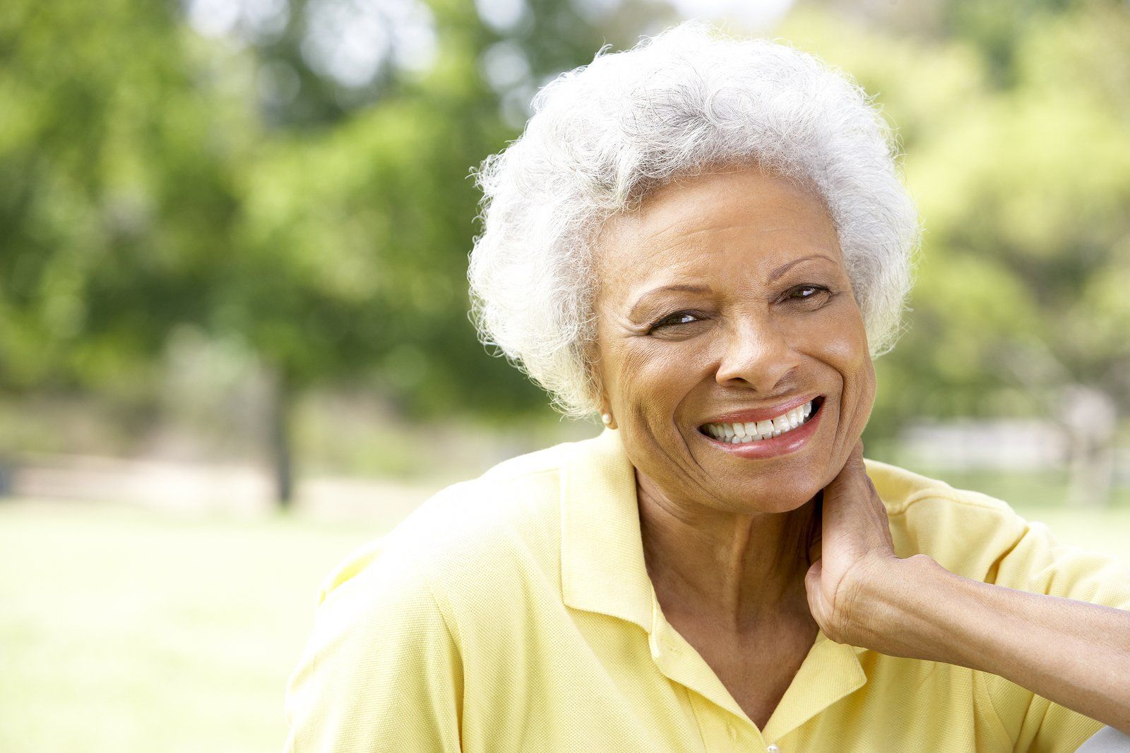 An elderly woman in a yellow shirt is smiling in a park.