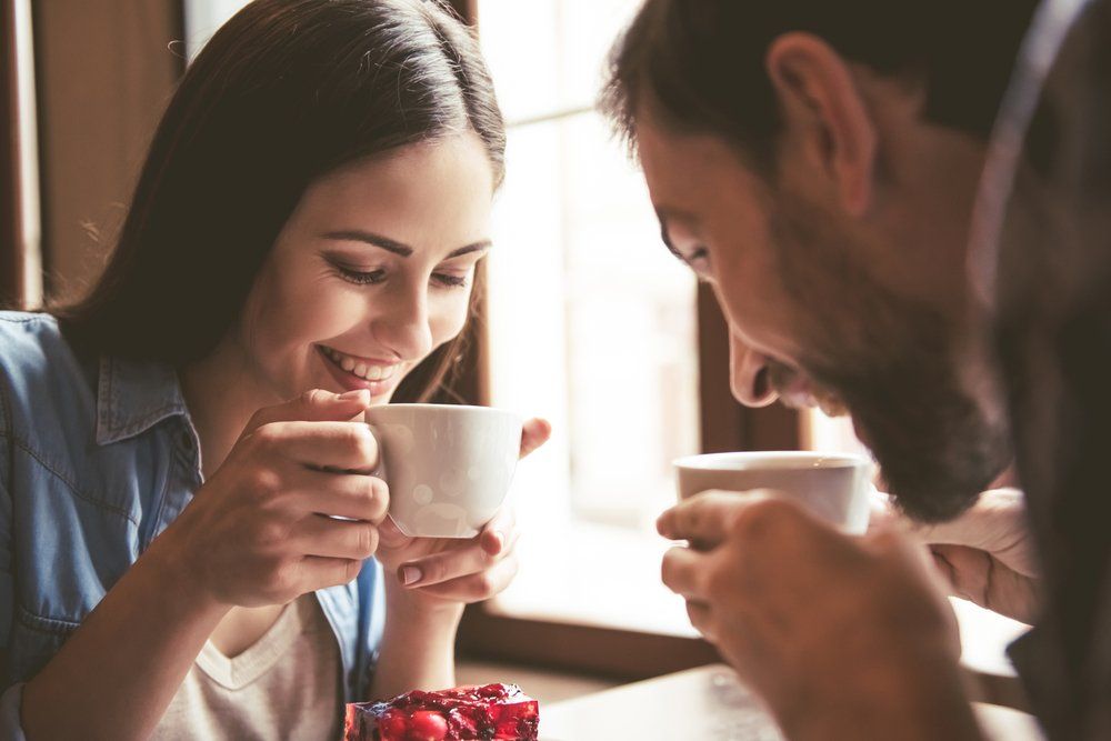 Zwei lächelnde Menschen sitzen an einem Tisch am Fenster und trinken Kaffee.
