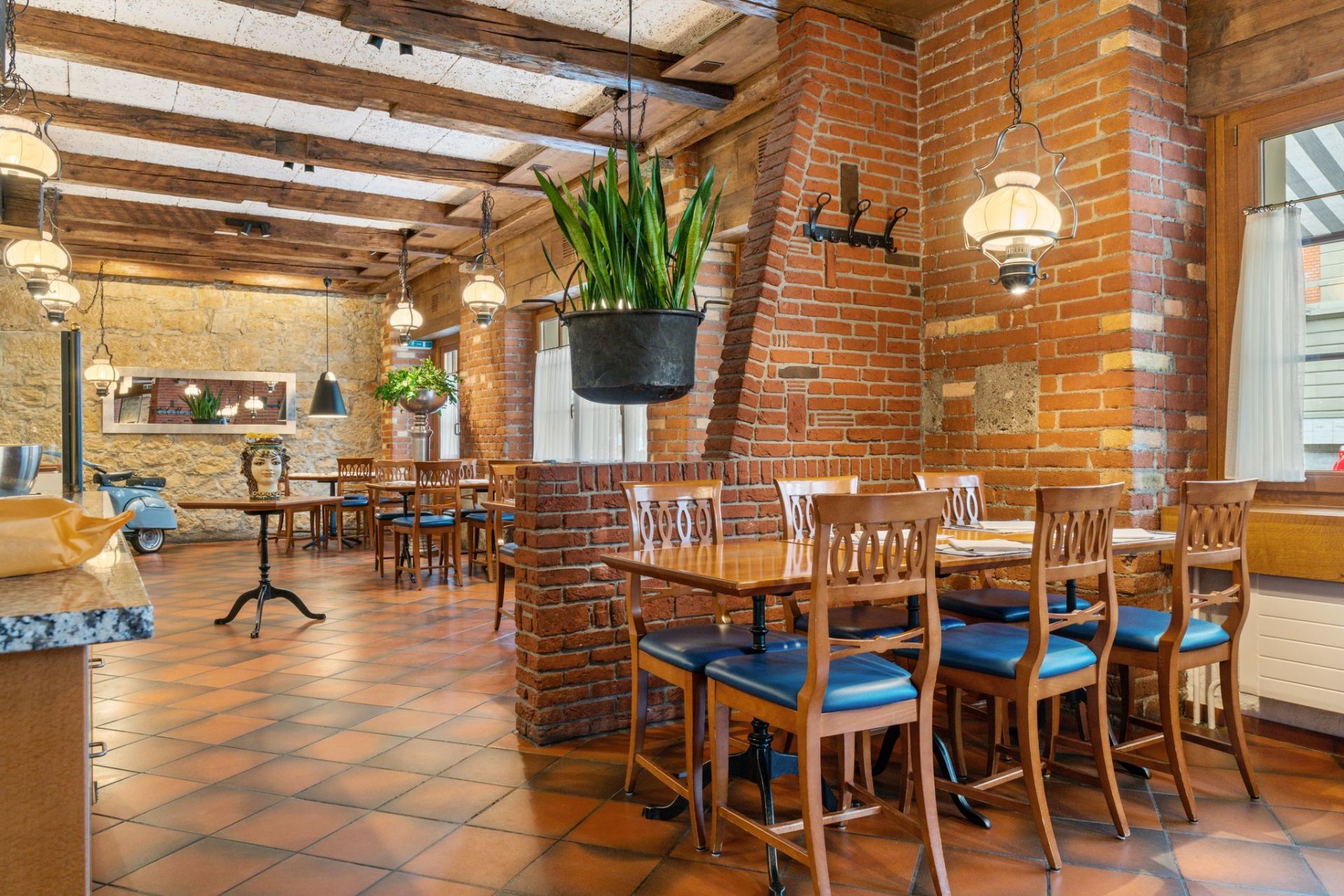 Rustikales Restaurantinterieur aus Backstein mit Holztischen, blauen Stühlen und Hängepflanzen