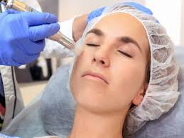 Una mano enguantada aplica un tratamiento facial con una jeringa a una persona recostada en un salón de belleza.