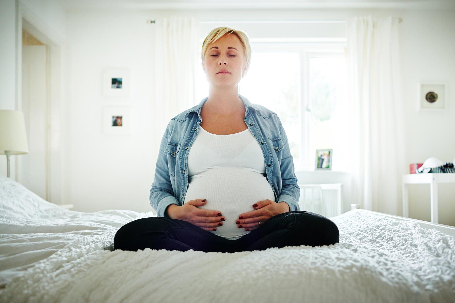 A pregnant woman is sitting on a bed with her eyes closed.