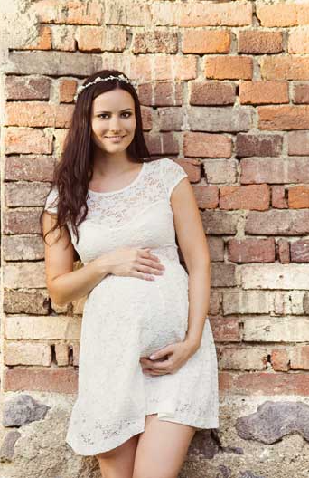 A pregnant woman in a white dress is standing in front of a brick wall.