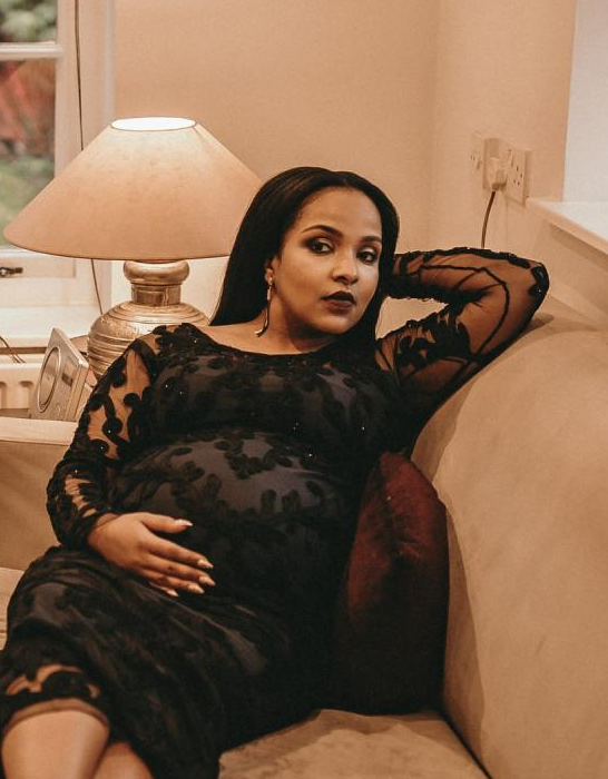 A pregnant woman in a black dress is sitting on a couch holding her belly.