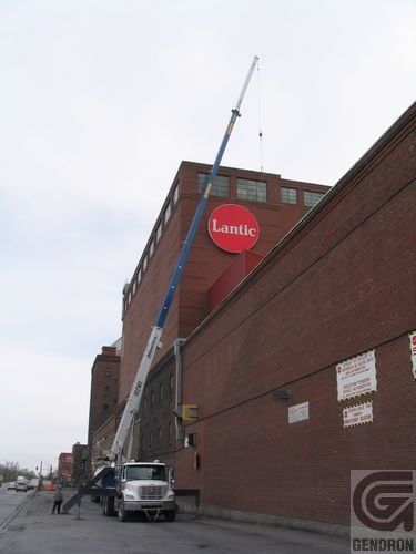 Grue déployée au-dessus d'un bâtiment portant l'enseigne « Lustig ». Camion dans la rue. Bâtiment en briques.