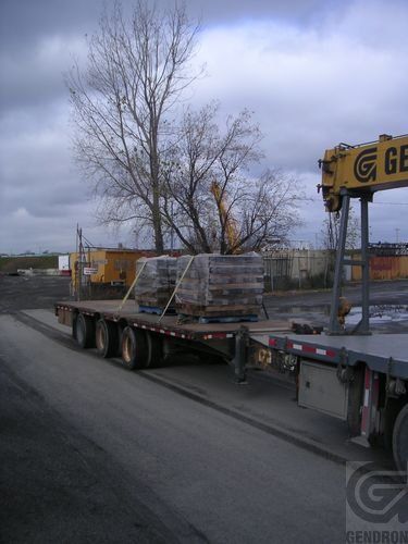 Camion plateau transportant des palettes filmées à l'extérieur. Une grue est visible à droite, ciel nuageux.
