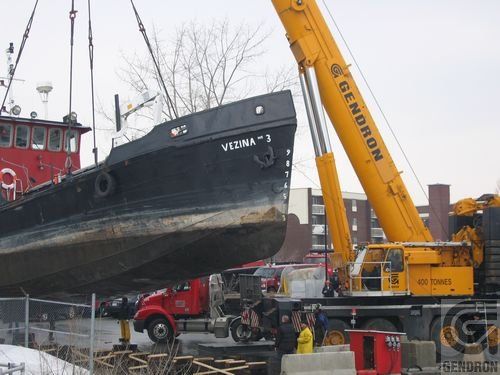 Le navire noir « Vizna 3 » est soulevé par une grue jaune sur le front de mer d'une ville.