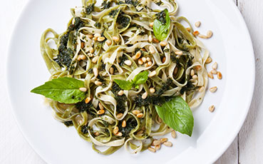 Fettuccine pasta with pesto, pine nuts, and basil leaves on a white plate.