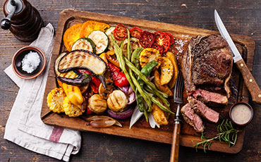 Steak with grilled vegetables on a wooden platter with salt, pepper, and dipping sauce.