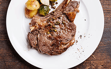 Steak with peppercorns on white plate with roasted potatoes and vegetables.
