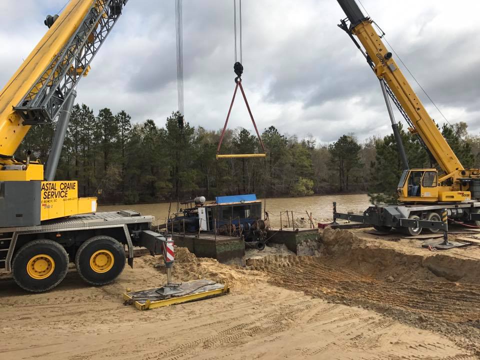 Two yellow cranes lifting a beam at a muddy construction site