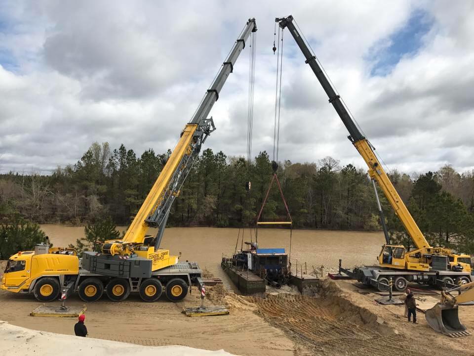 Two yellow cranes lifting a large object at a muddy construction site near a wooded field