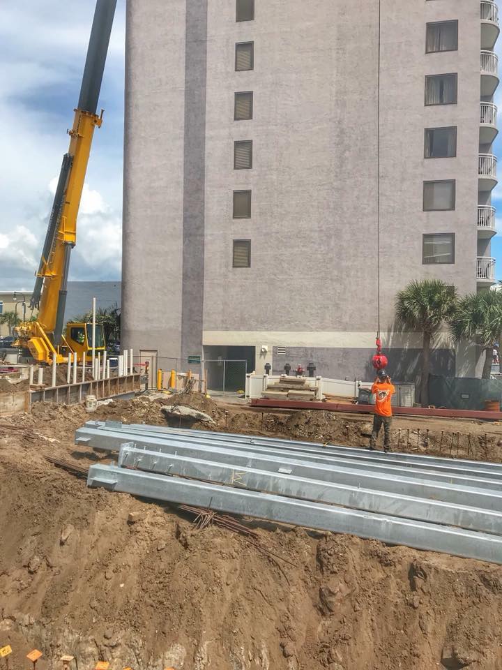 Construction site beside a tall building, with a crane, workers, and stacked metal beams in a dug trench.
