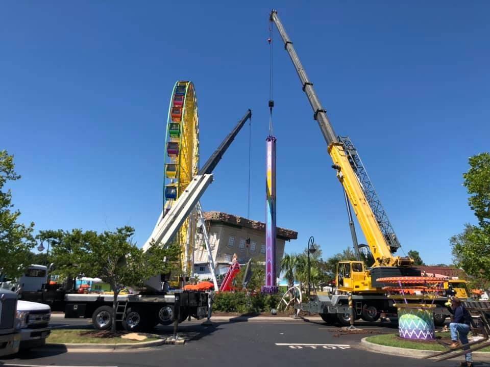 Two cranes assembling a tall Ferris wheel at an outdoor fairground under a blue sky