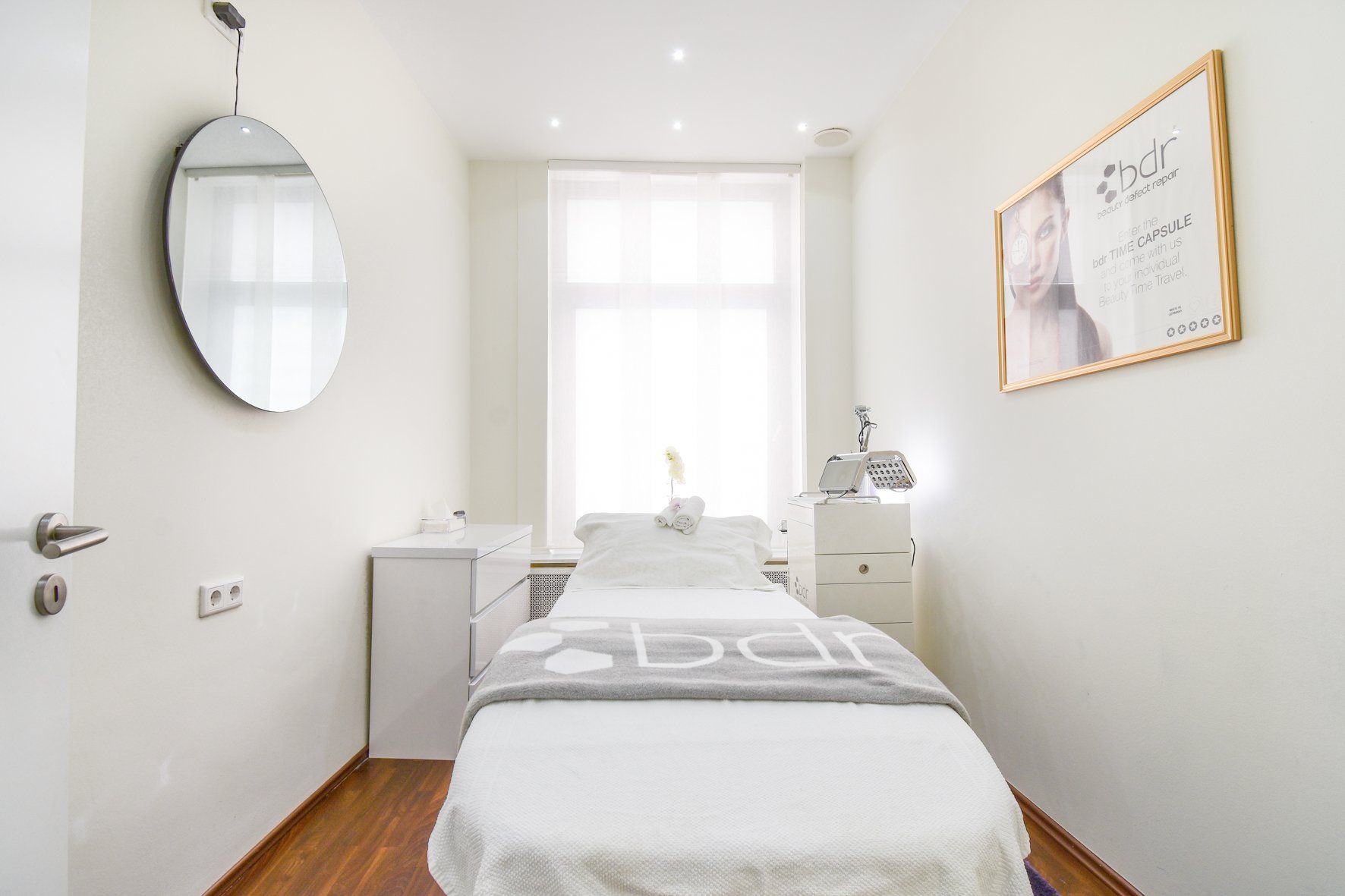 Ein heller, minimalistischer Spa-Raum mit Behandlungsliege, Spiegel und Fenster. Weiße Wände und Holzböden sorgen für eine klare Ästhetik.