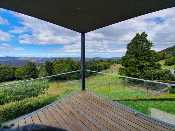Wooden deck with glass railing overlooking green fields, trees, and blue sky.