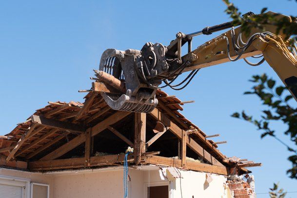 Picture of an excavator machine clamped onto the roof of a building pulling the roof off.