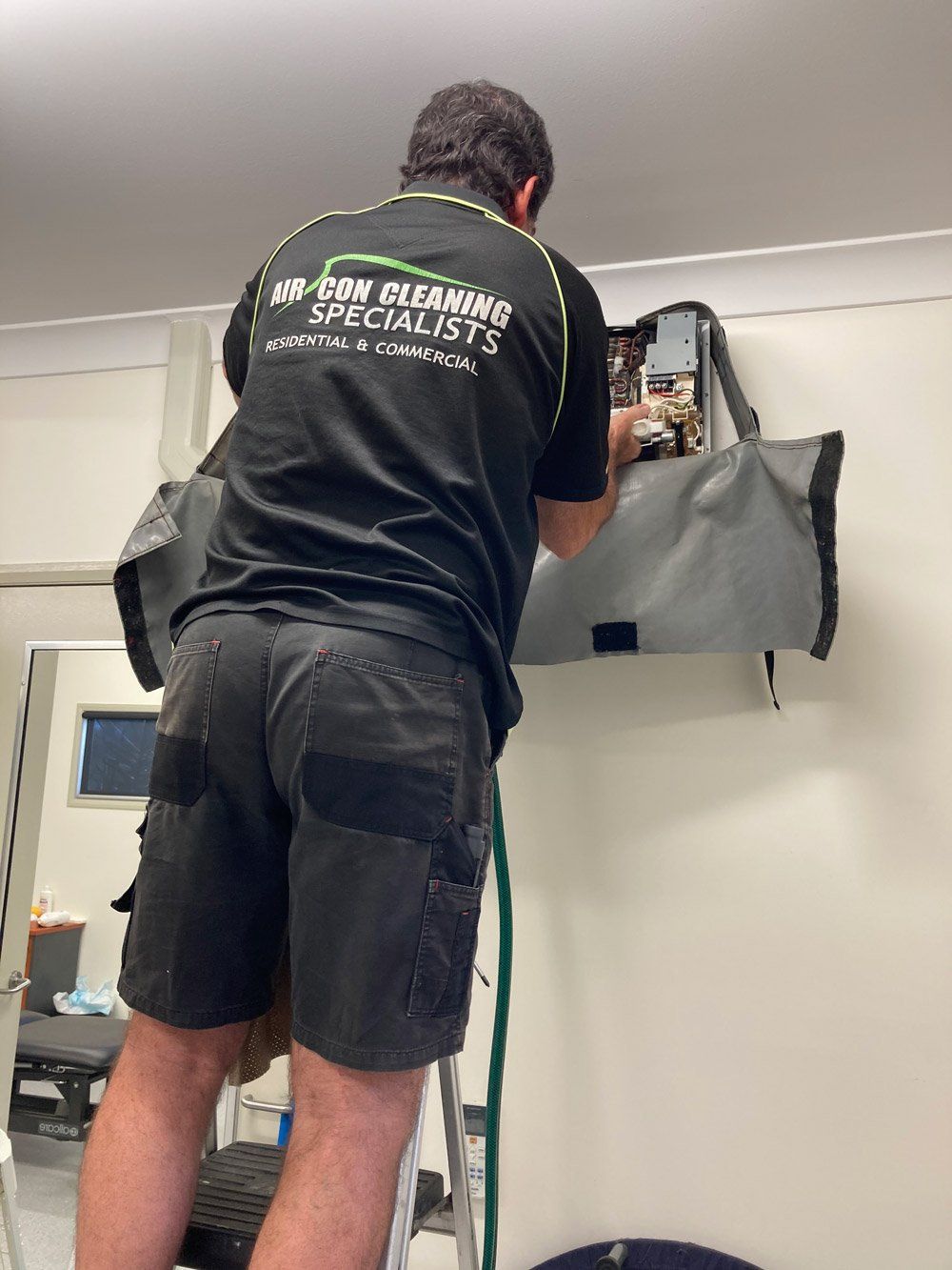 A Man is Standing on a Ladder Working on an Air Conditioner — Air-Con Cleaning Specialists in Redlynch, QLD A Man is Standing on a Ladder Working on an Air Conditioner — Air-Con Cleaning Specialists in Redlynch, QLD