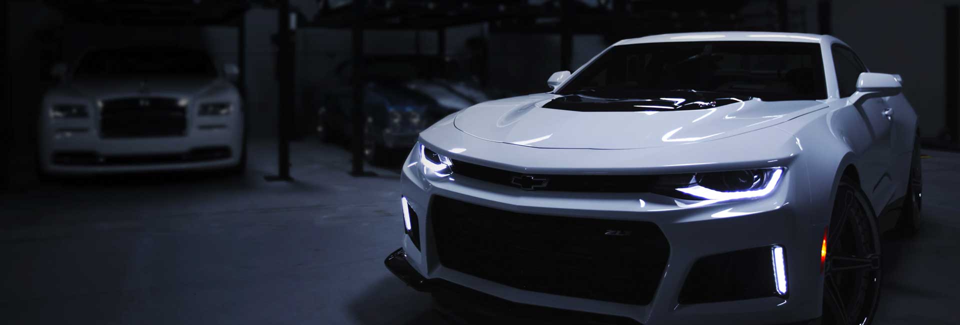 A white car is parked in a dark garage next to another car.