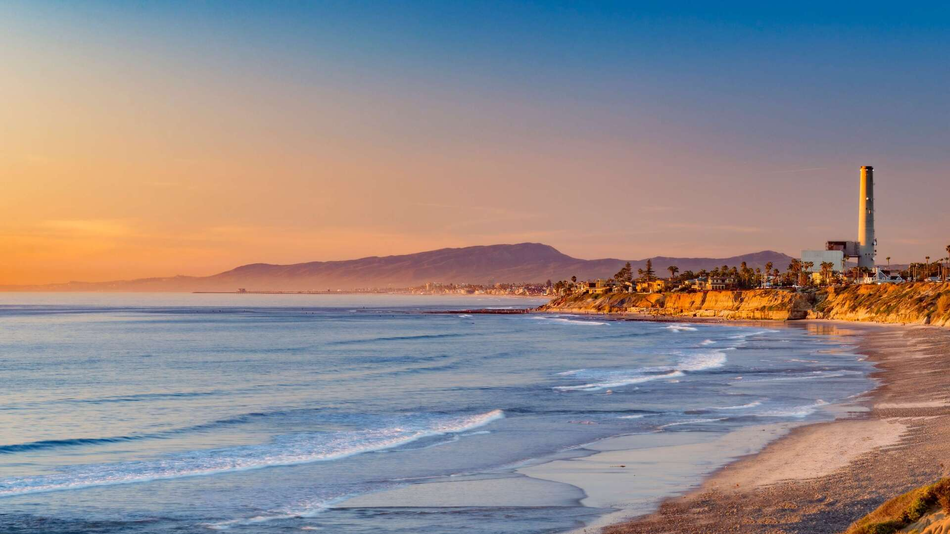 Beach scene at sunset, ocean waves rolling onto the shore, with mountains and a tall smokestack in the distance.