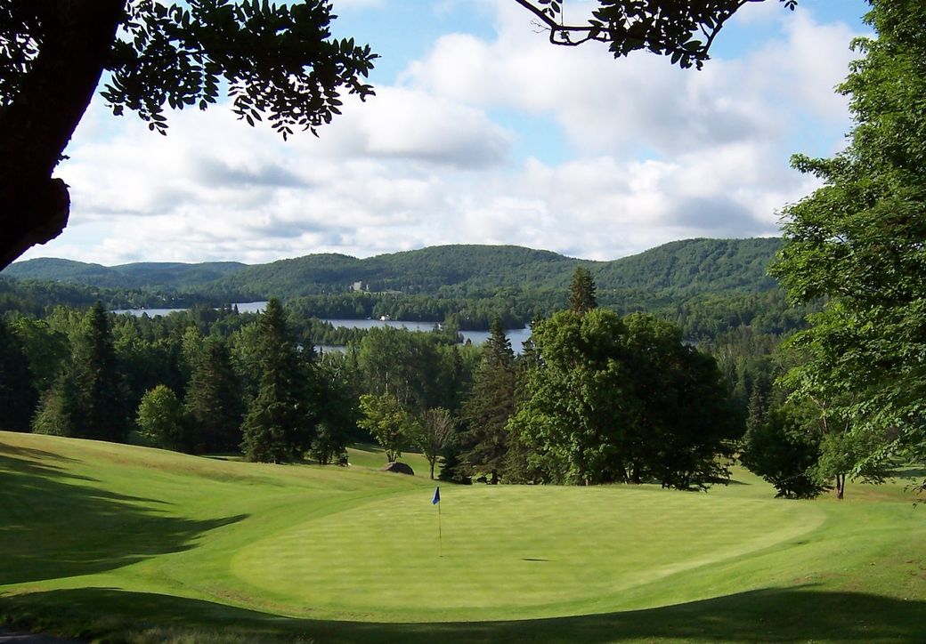 Green golf course with a view of a lake and forested hills under a blue sky with white clouds.